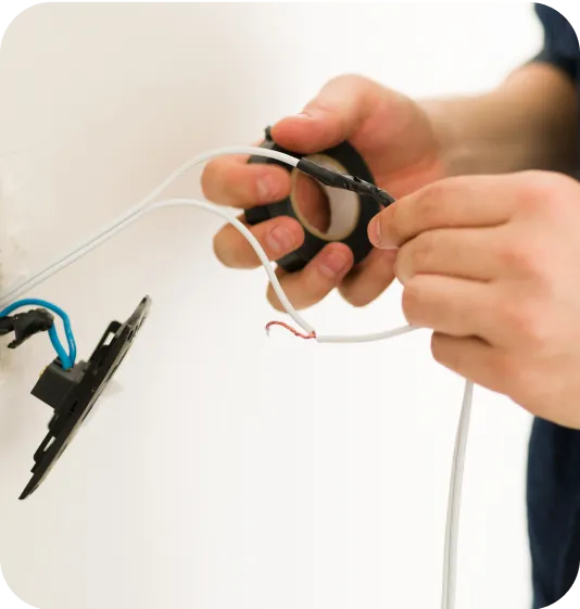 Hands wrapping black electrical tape around exposed wires near a wall outlet.