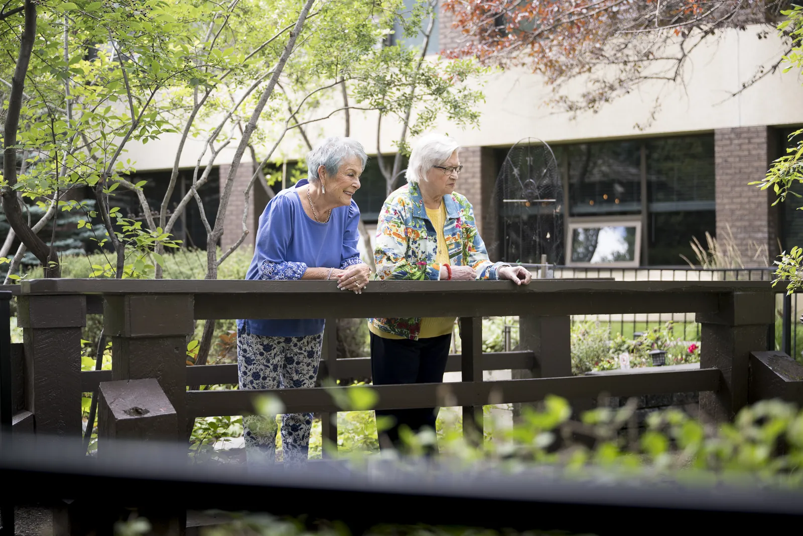 Two smiling residents lean on the railing of the wooden bridge in a leafy Trinity Lodge courtyard