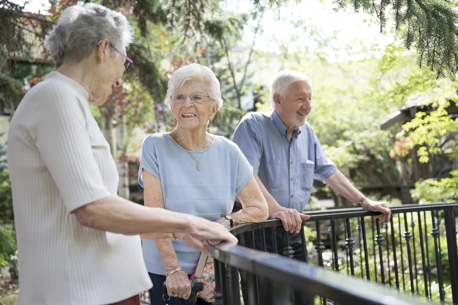 Three residents chat by a railing in a sunny Trinity Lodge courtyard