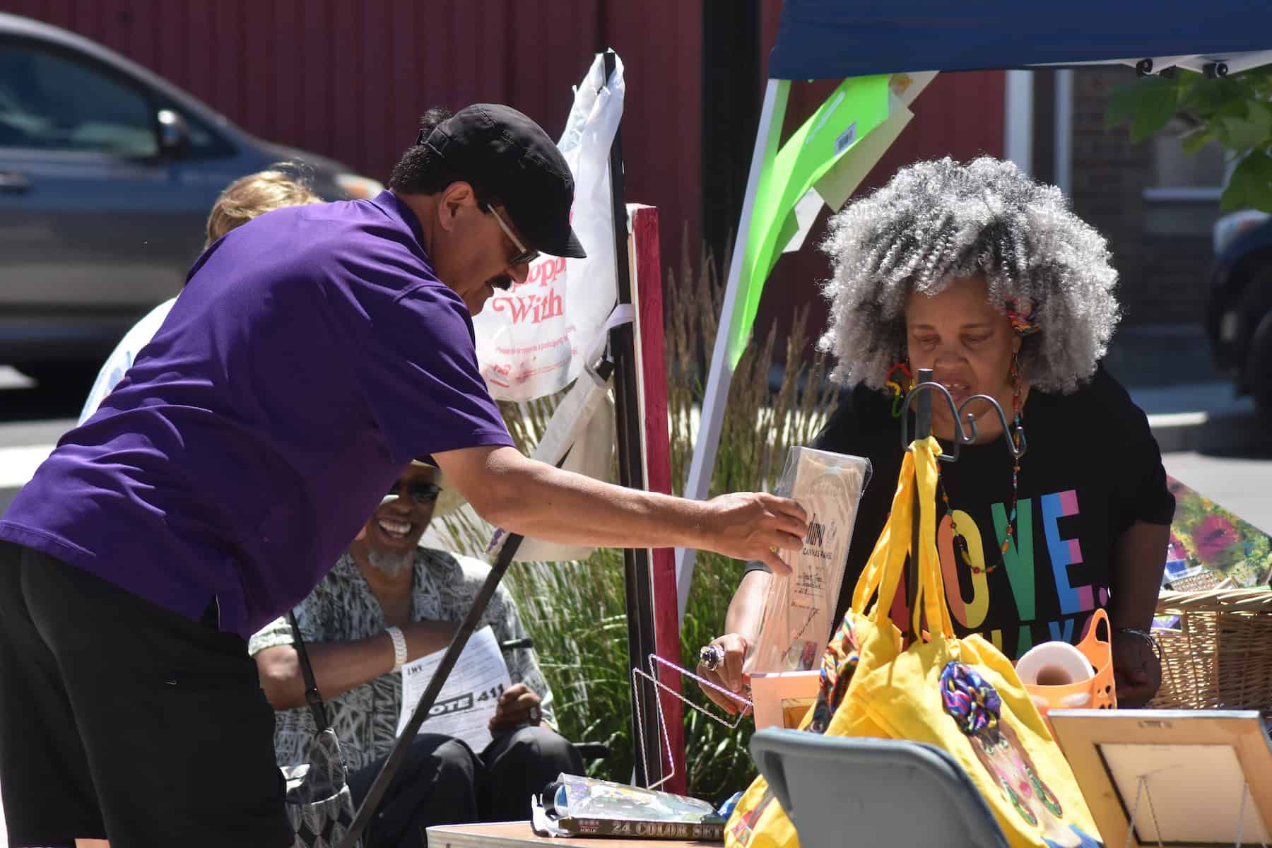 A man in a purple shirt leans over an outdoor vendor table to examine artwork while a woman in a black "MOVE" shirt looks on, surrounded by colorful tote bags and art supplies at a community event.