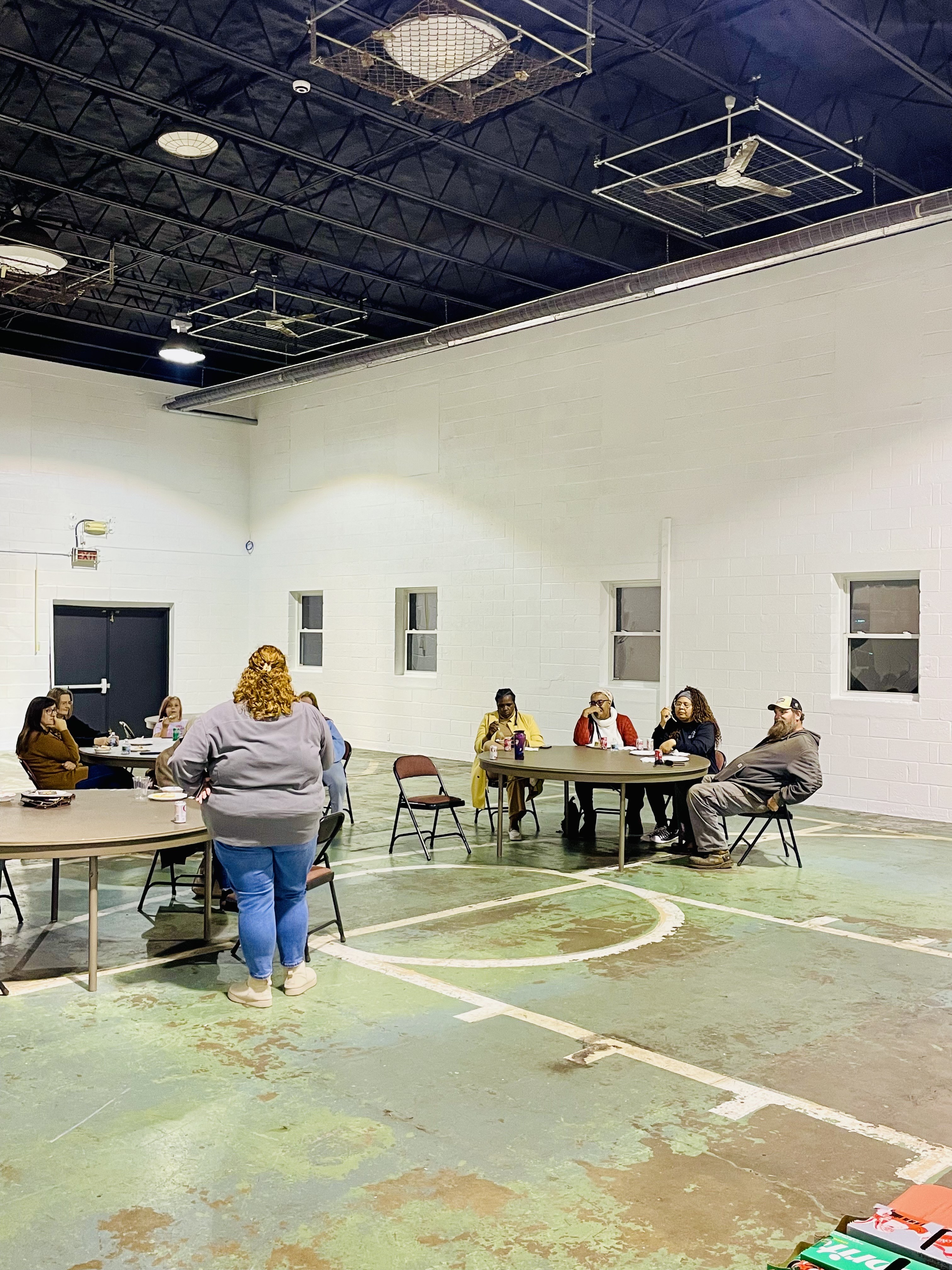 A diverse group of people gathered in a school gymnasium for a community-building event hosted by Lightkeepers, with banners and interactive stations in the background.