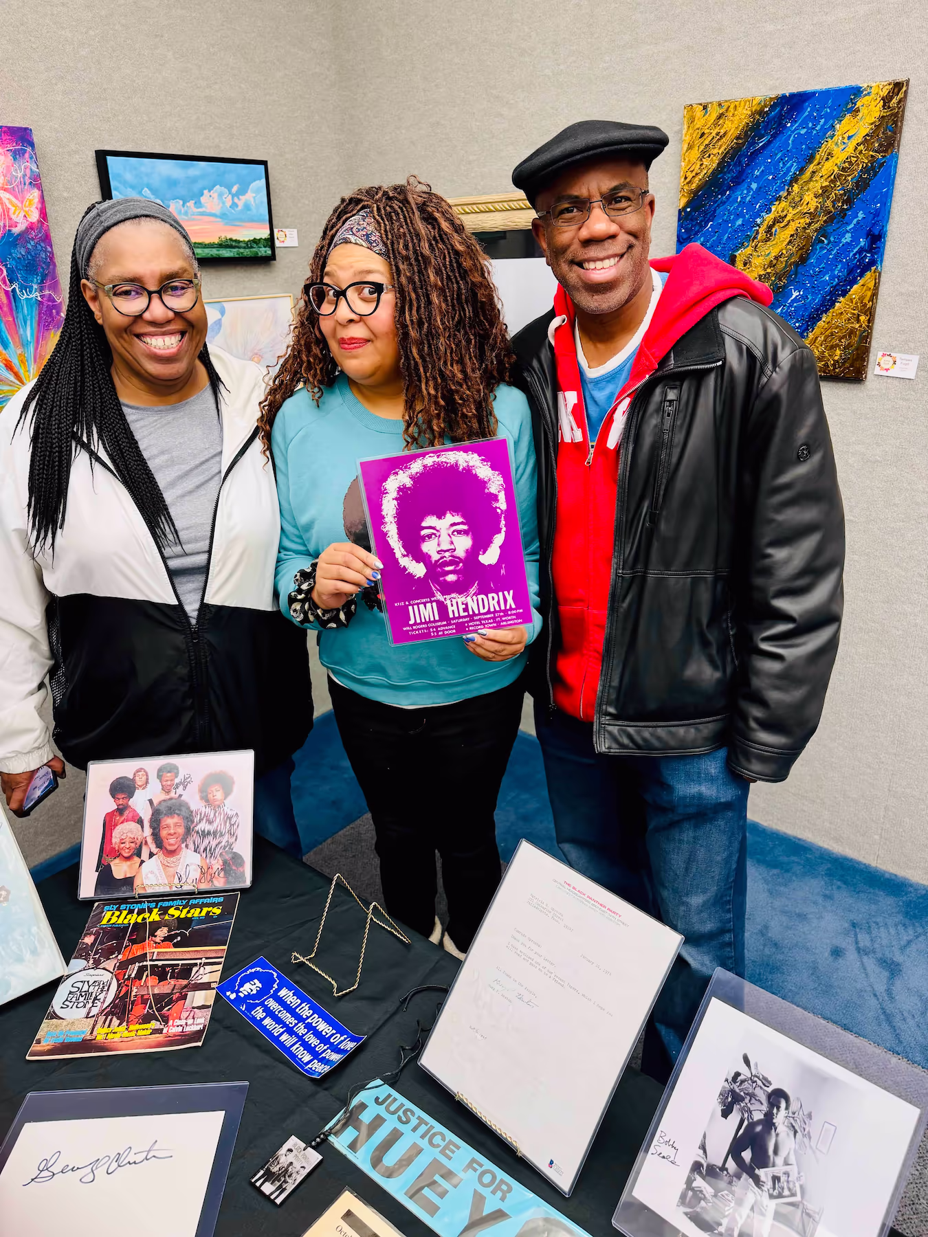 Three people smile at the camera while standing behind a table filled with Black history memorabilia, including a Jimi Hendrix poster.