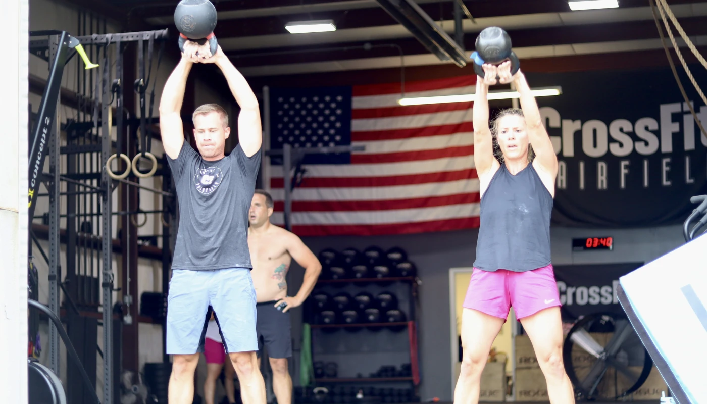 A man and a woman perform kettlebell exercises in a CrossFit gym, with an American flag visible in the background.