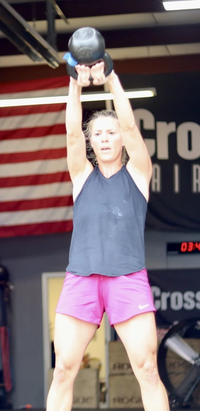 A man and a woman perform kettlebell exercises in a CrossFit gym, with an American flag visible in the background.
