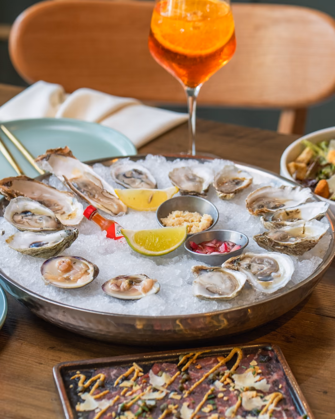 Metal tray filled with crushed ice holding fresh oysters on the half shell around lemon wedges, a small bottle of hot sauce, and metal cups with condiments, set on a wooden table with a glass of orange cocktail and plates in the background.