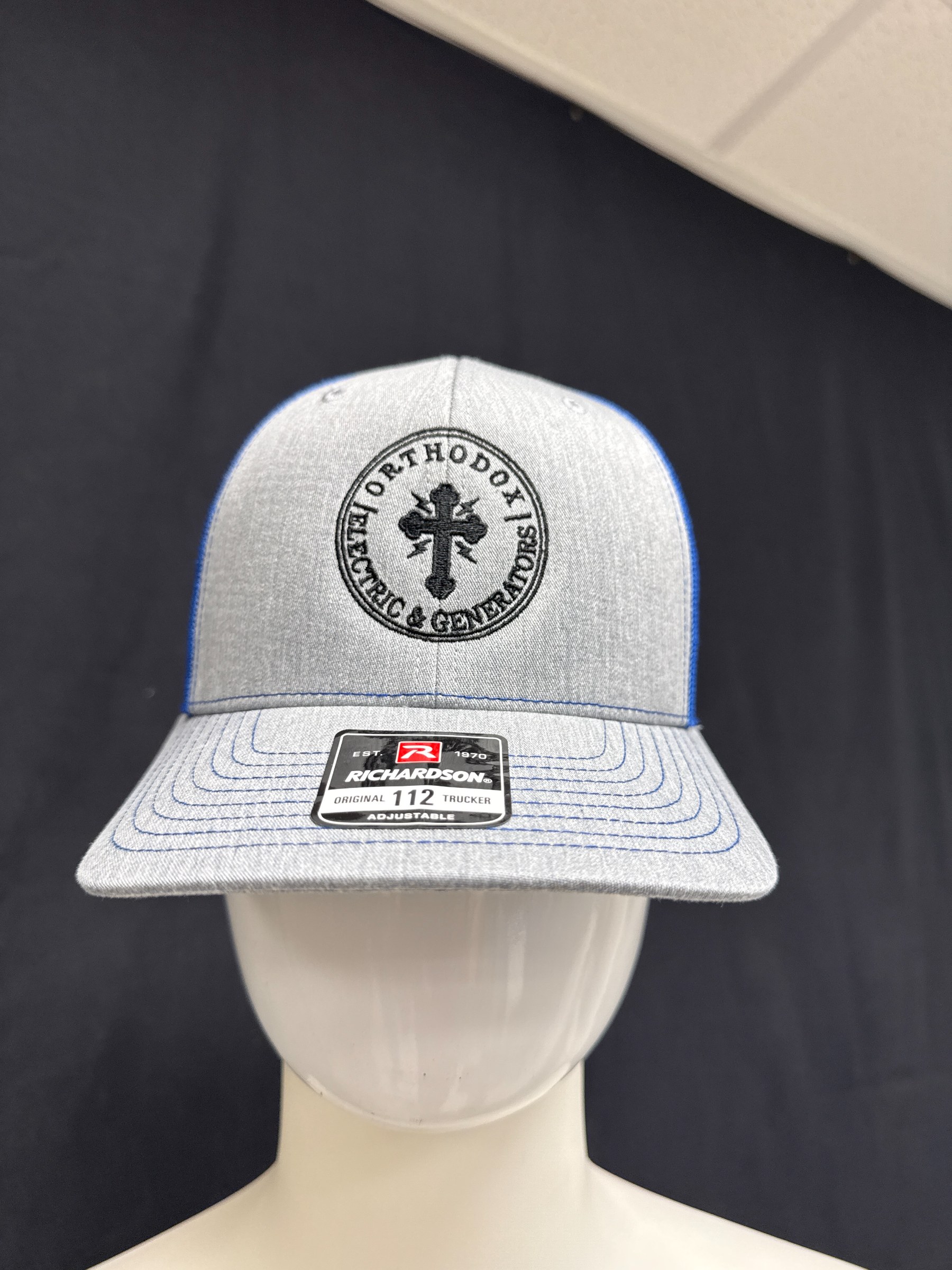 Gray Richardson Original 112 trucker cap with blue stitching and mesh back on a mannequin, featuring a black embroidered circular logo with a cross and the text 'Orthodox Electric & Generators'.