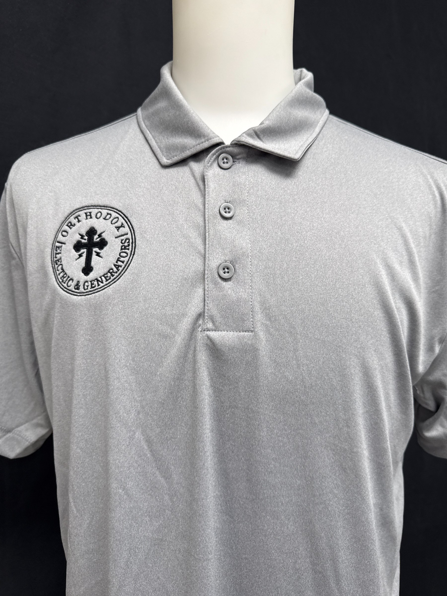 Light gray polo shirt with three buttons and an embroidered black logo on the left chest featuring a cross and the text 'Orthodox Electric & Generators'.