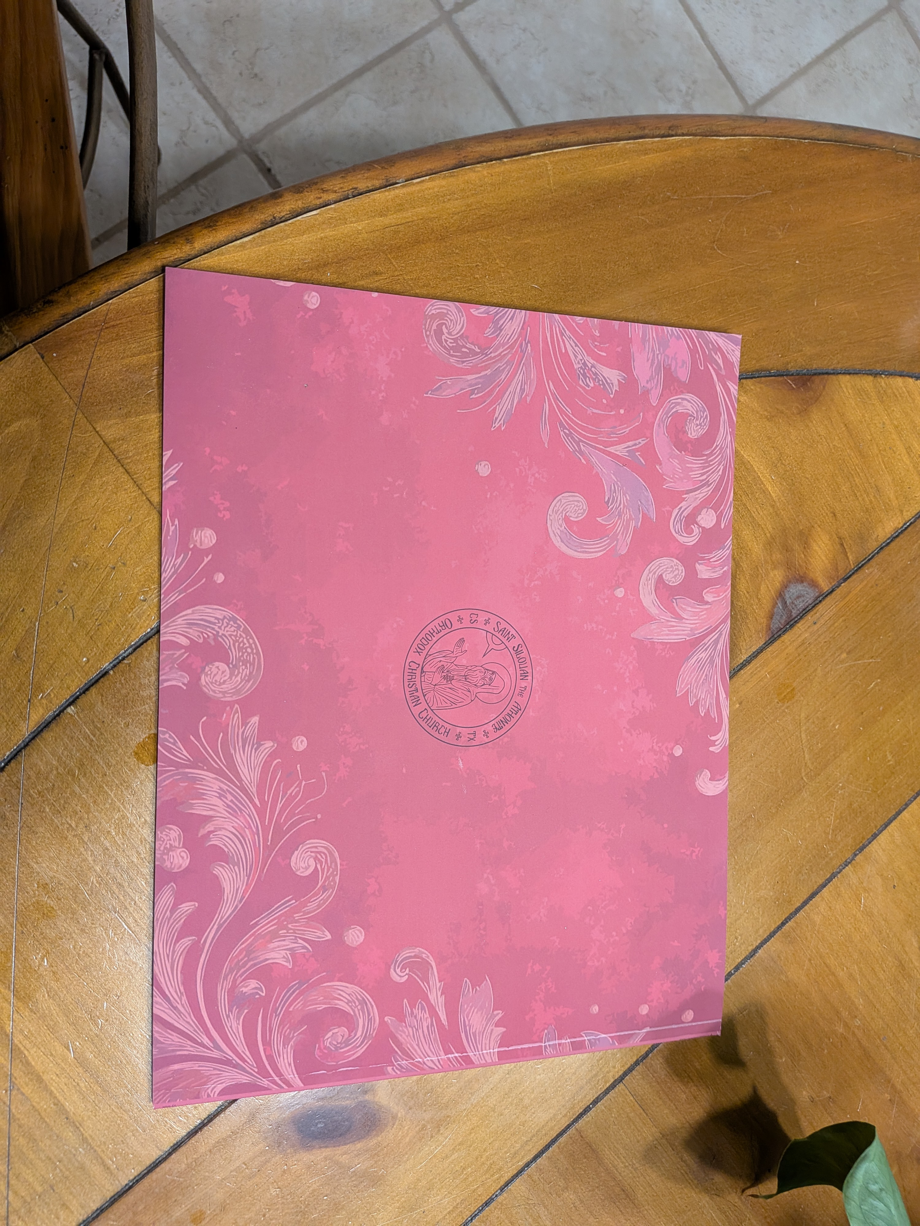 Closed book or booklet with a pink cover featuring elegant light pink floral swirls and a small circular emblem with text and an illustration in the center, placed on a wooden table with beige tiled floor in background.