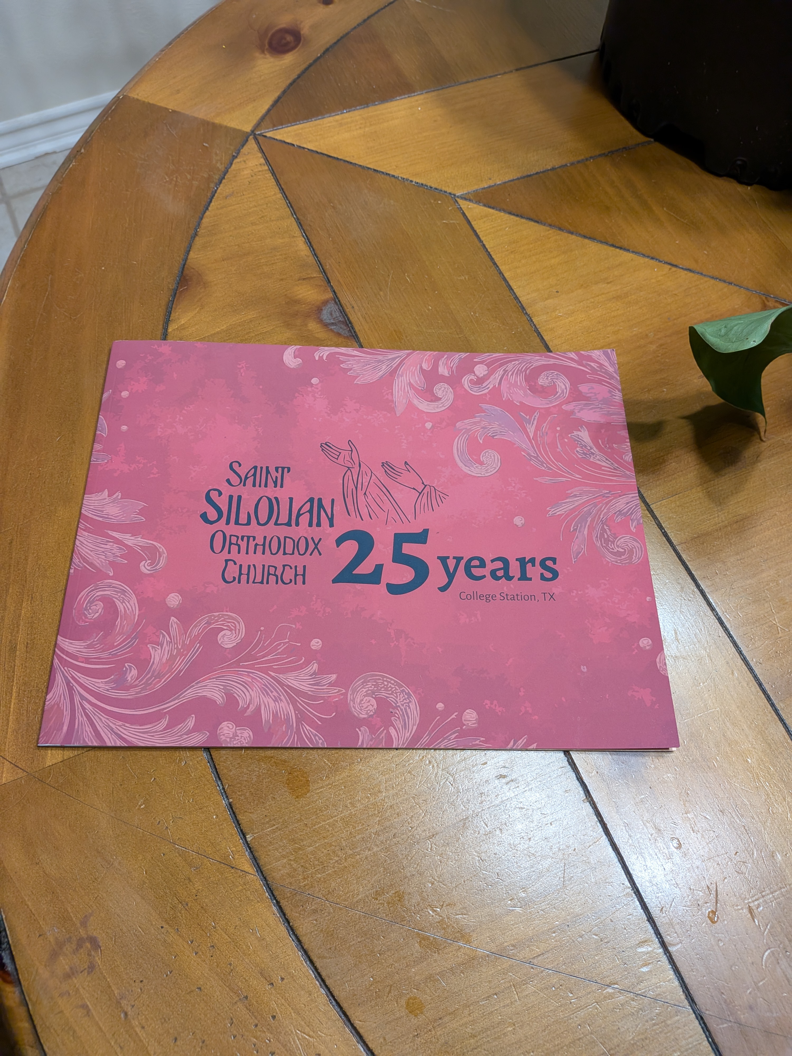 A reddish booklet cover on a wooden table with ornate floral patterns and text reading 'Saint Silouan Orthodox Church 25 years College Station, TX' with an illustration of two raised hands in the center.