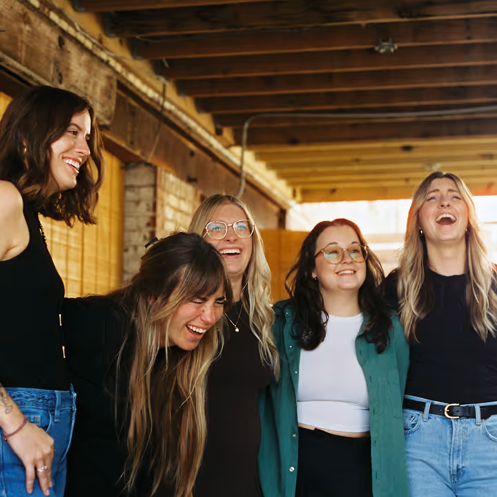 A group of five women laughing and hugging under a wooden roof, clearly enjoying the moment together.