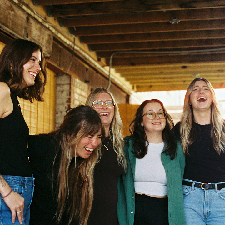A group of five women laughing and hugging under a wooden roof, clearly enjoying the moment together.
