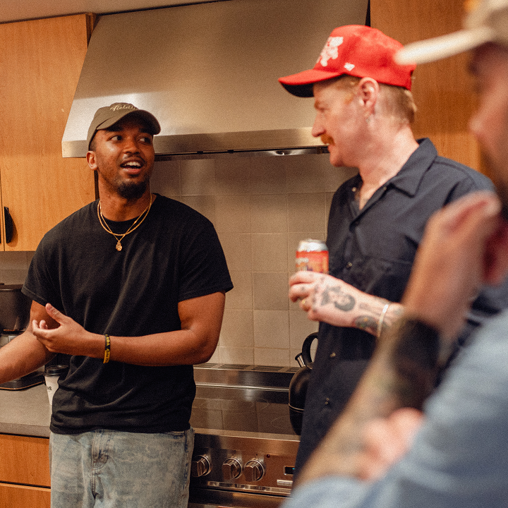 Two men having a casual conversation in the kitchen while holding drinks, with a third person listening in.