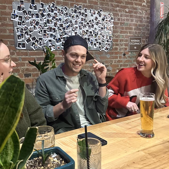 Three people sitting at a bar table, smiling and enjoying drinks with a photo collage in the background.