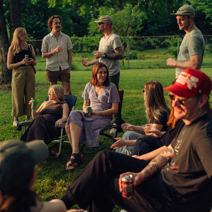 A group of colleagues gathered outside, sitting and standing in a circle with drinks in hand, enjoying a casual get-together.