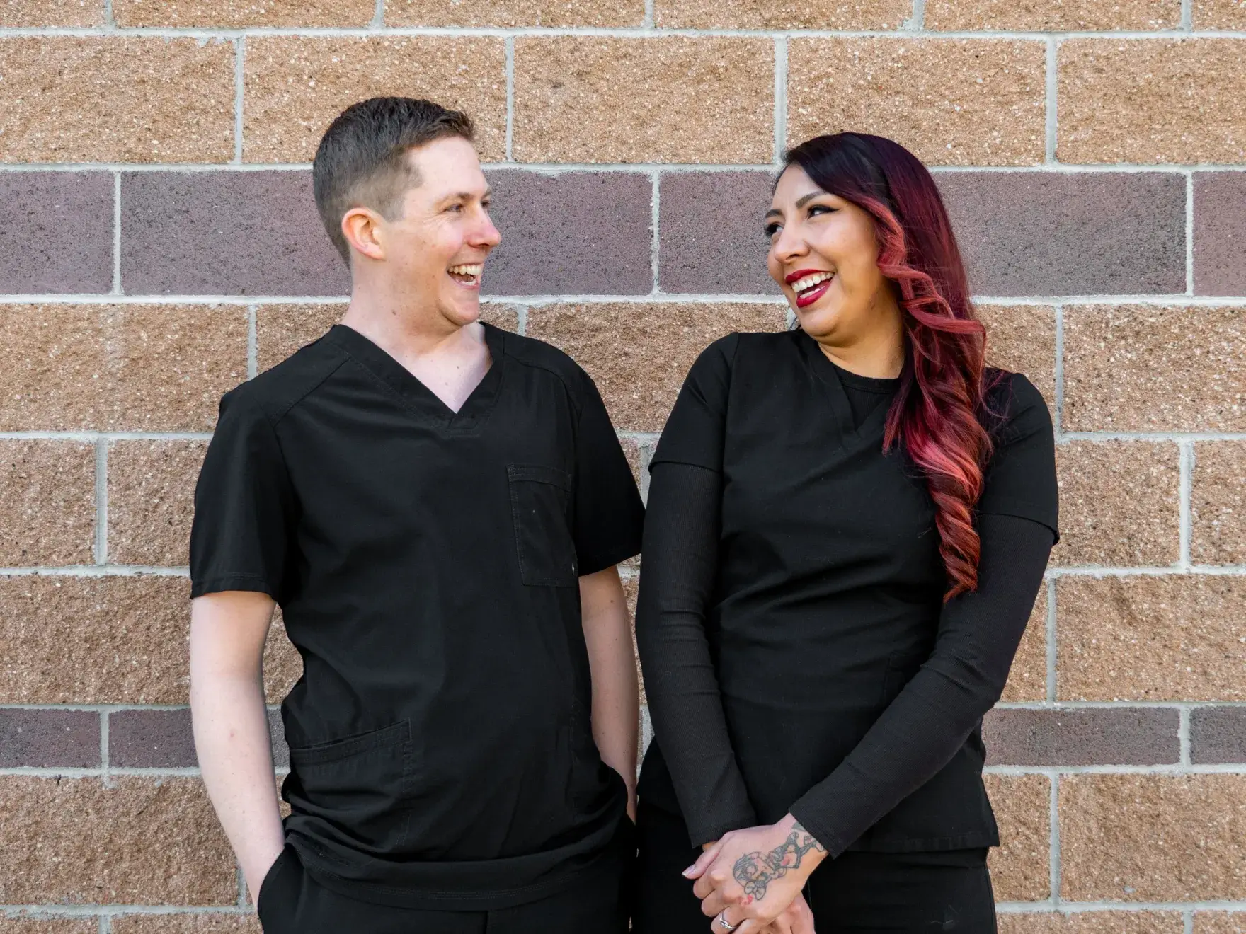 Two people in black scrubs stand against a brick wall, smiling with arms crossed.