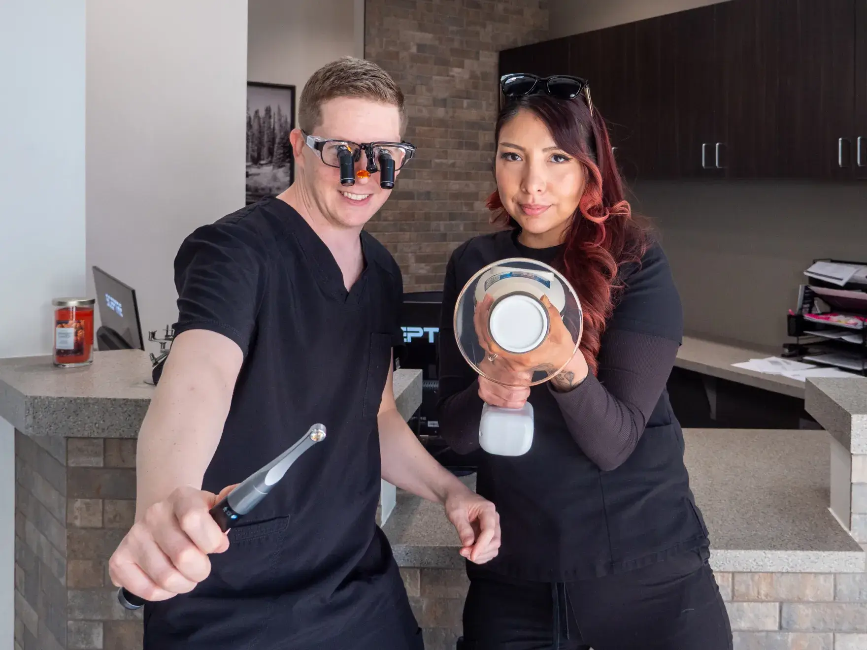 A woman in a dental chair holds a hand mirror, while a man in scrubs smiles at her.