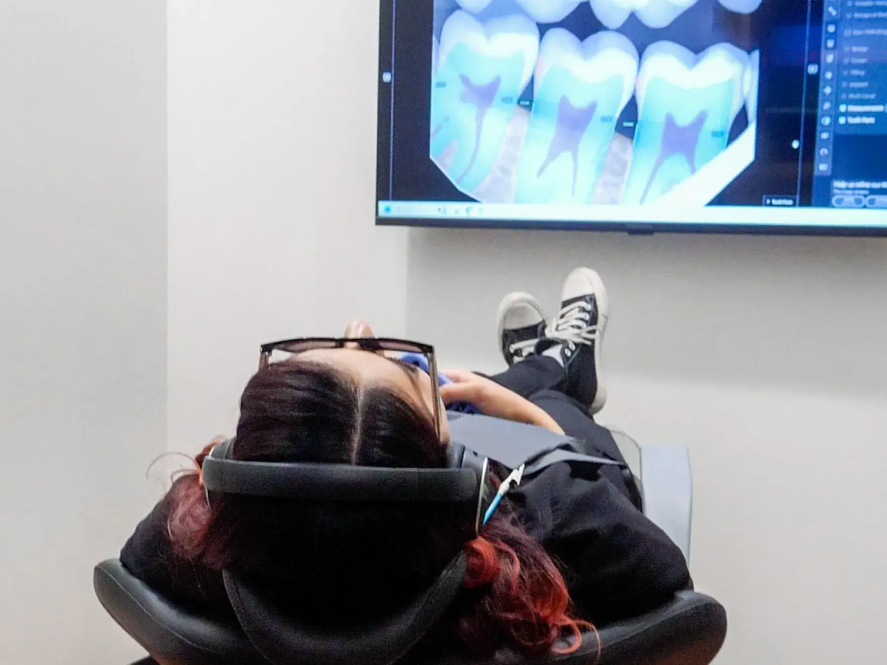 A woman holds a mirror, checking her teeth while dental X-rays are displayed on a screen.