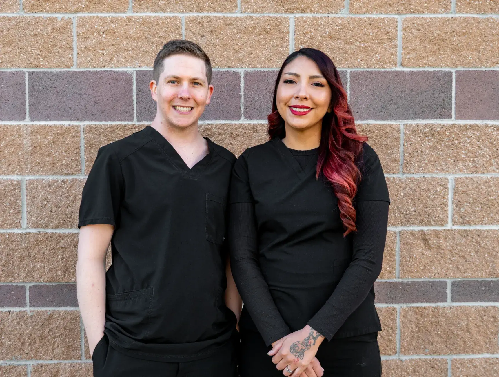 A person wearing black scrubs stands smiling with arms crossed in front of a brick wall.