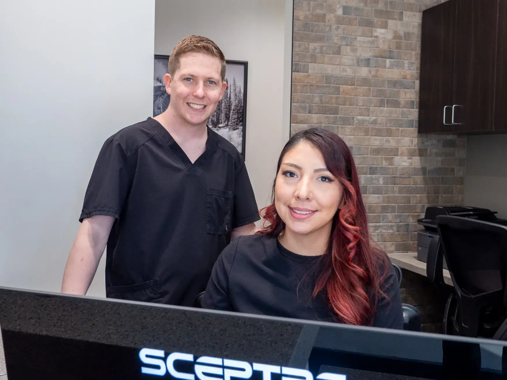Two people in black scrubs stand against a brick wall, smiling with arms crossed.