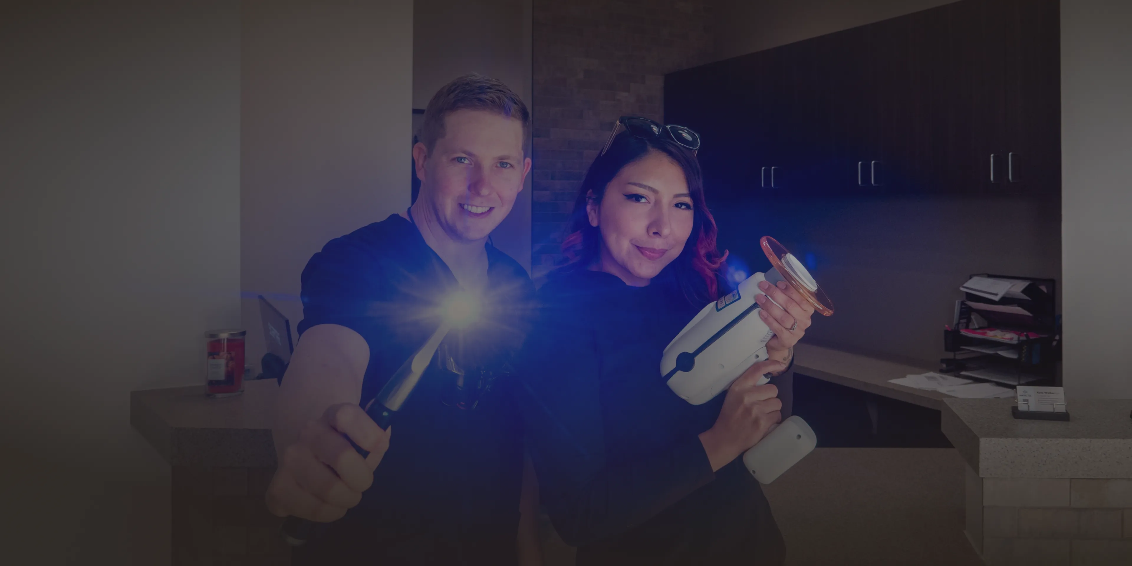 Two people smiling, holding dental equipment, with a bright light shining from one tool.