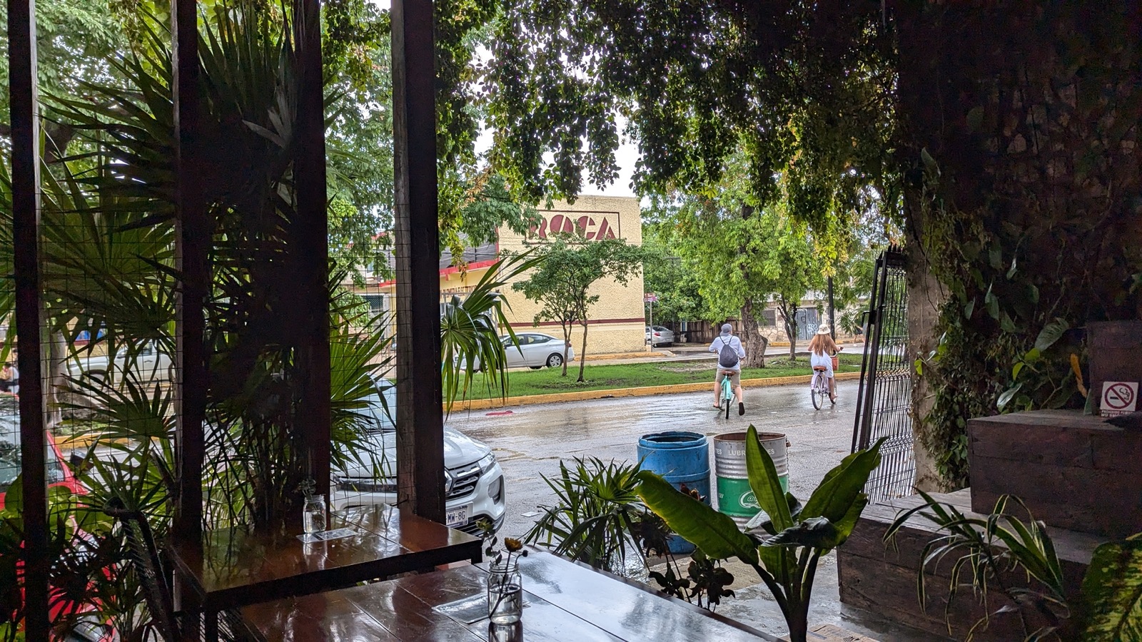 Looking out from inside a Tulum restaurant to the street, cyclists passing on a wet road