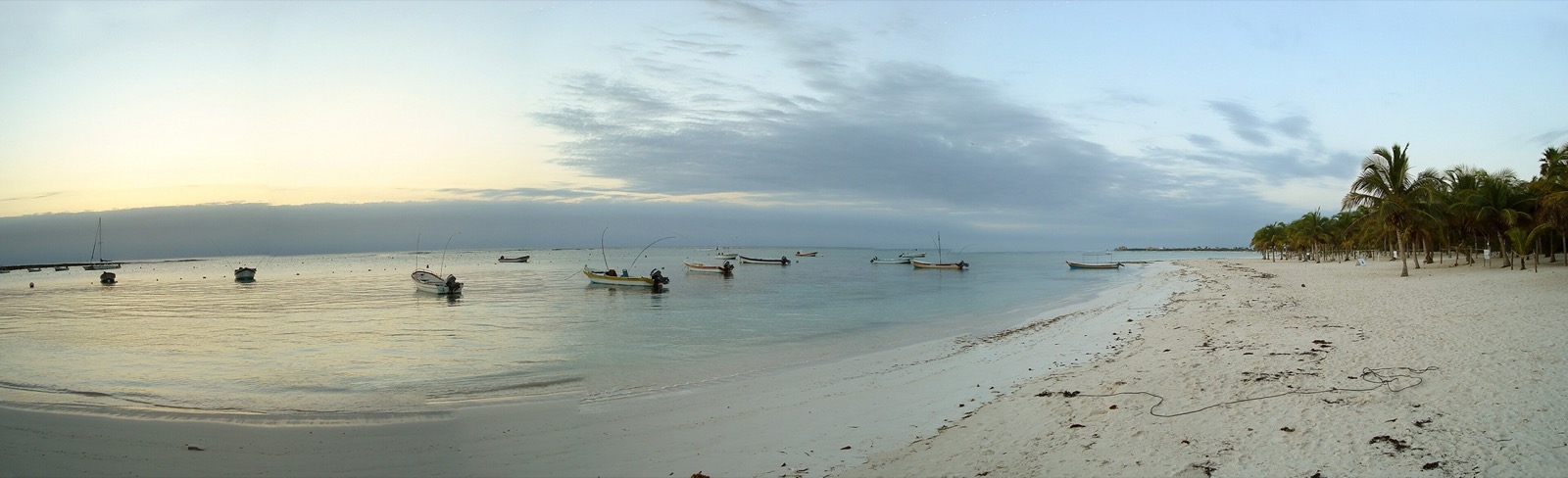 Panorama of Akumal Bay at dawn, with small boats moored on calm water and palms along a white sand beach