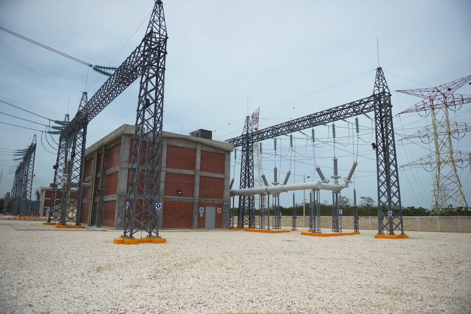 Steel gantries and a brick control building at the CFE Riviera Maya substation in Puerto Morelos, with low jungle beyond the perimeter wall