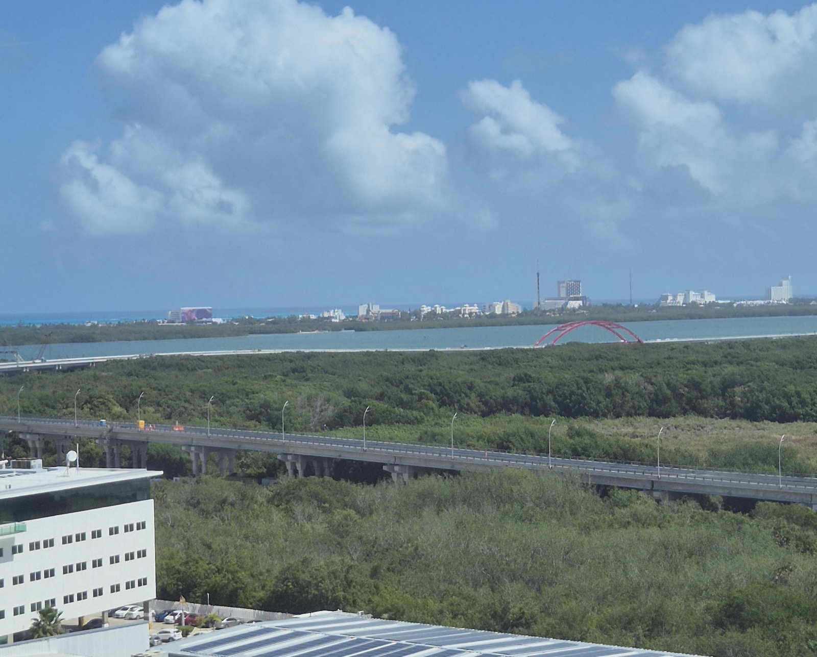 An elevated roadway over green mangrove with the red arch of the Nichupté bridge and hotel towers beyond