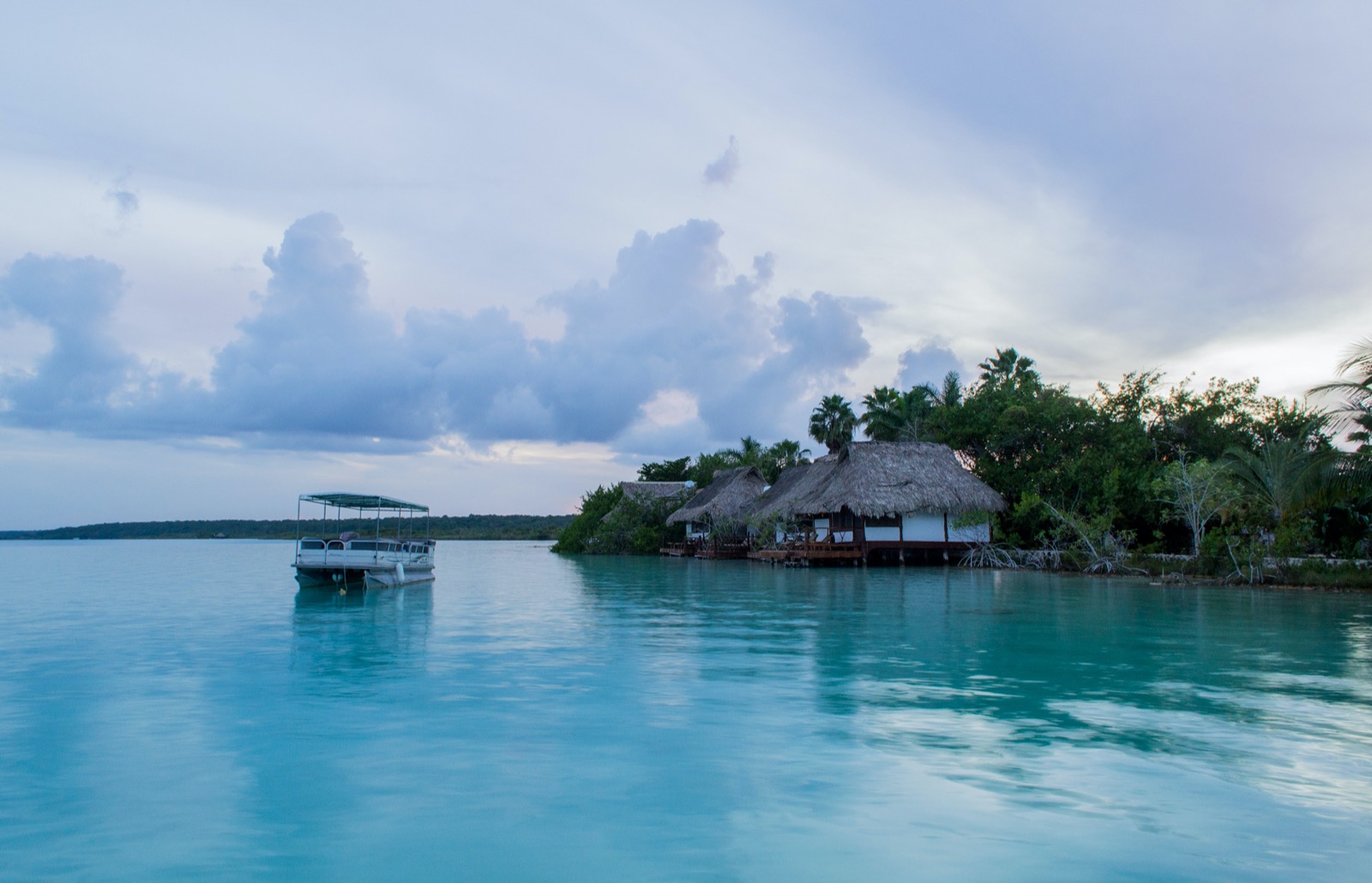 A tour boat moored on the turquoise water of the Bacalar lagoon beside thatched palapas at dusk