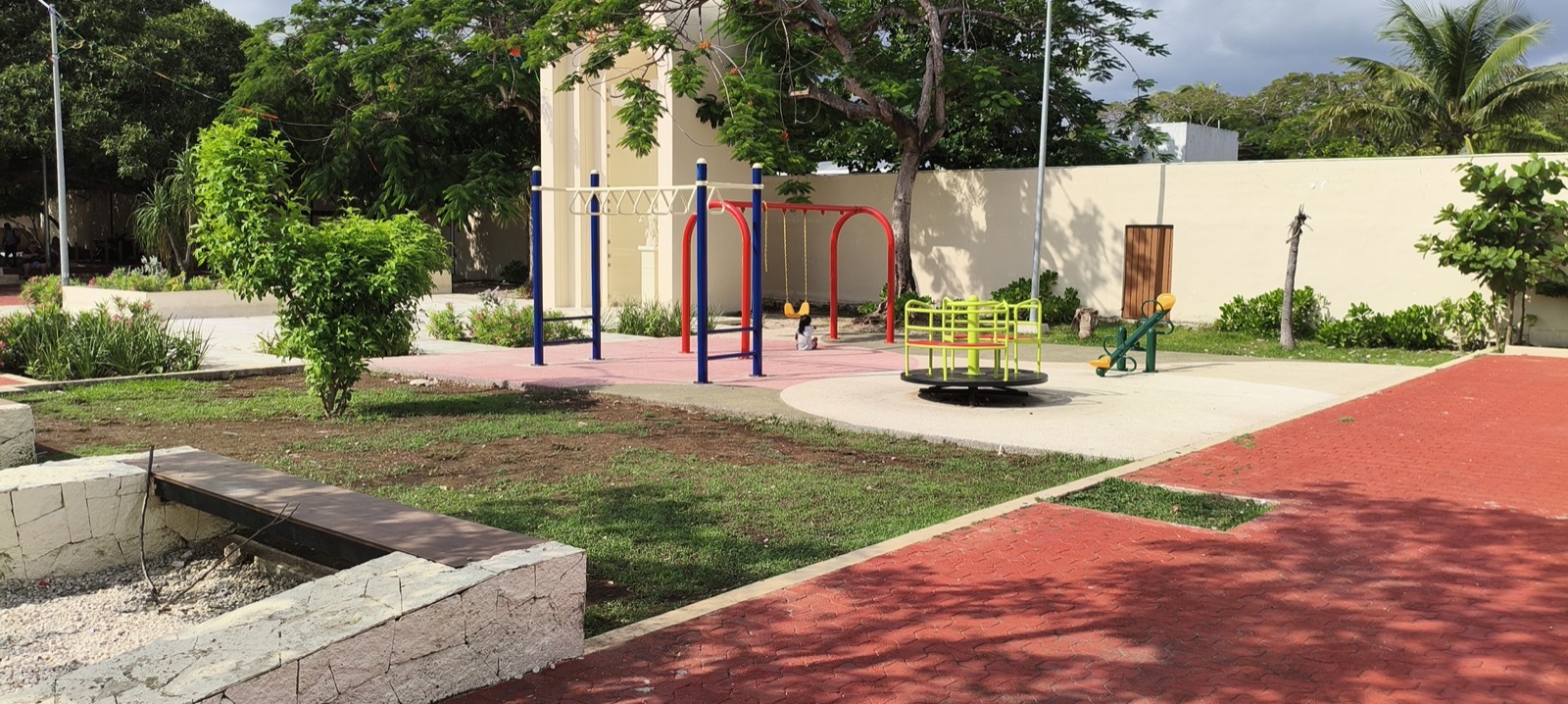 A playground with swings, climbing bars and a roundabout on a pink surface in Parque Leona Vicario, Playa del Carmen, edged by lawn, young shrubs and red interlocking paving