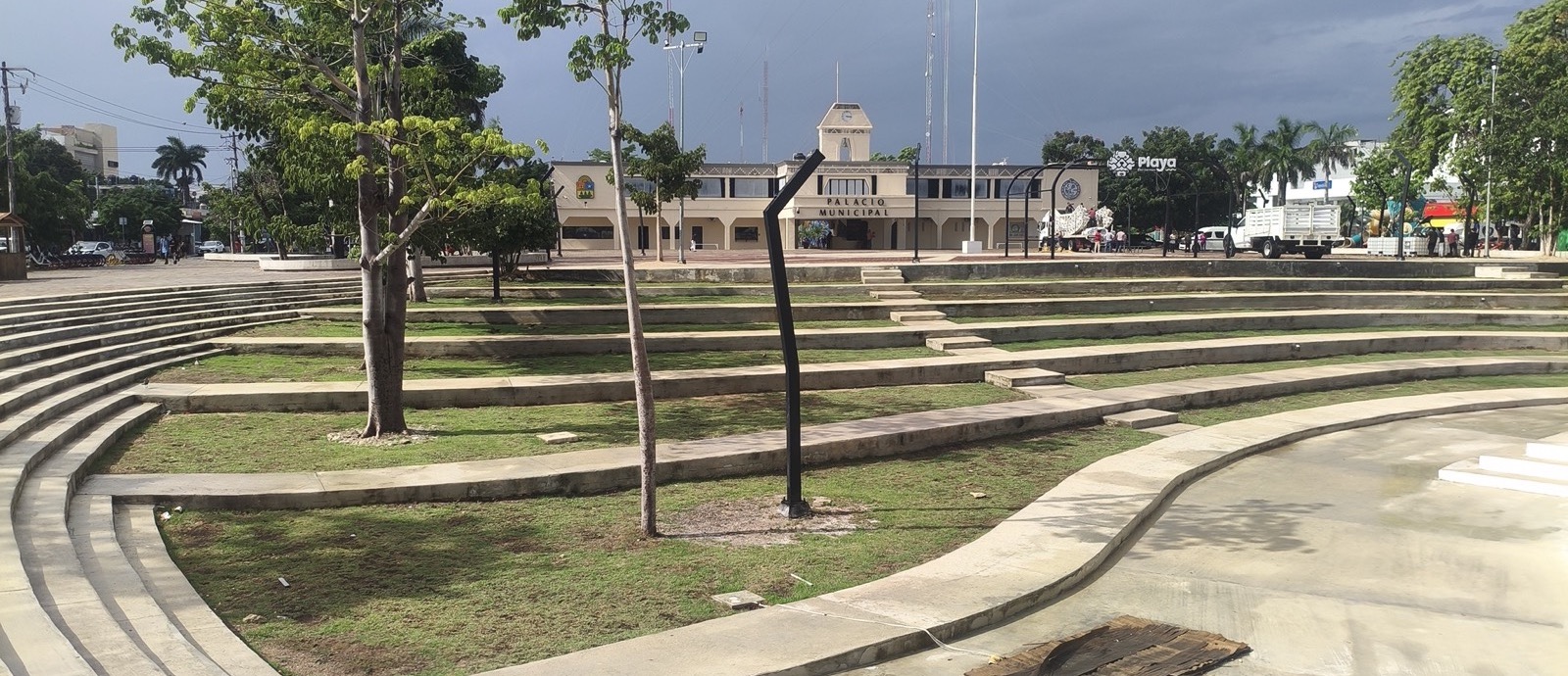 Terraced grass and concrete steps of the Plaza 28 de Julio amphitheater with young trees, and the cream colored Palacio Municipal of Playa del Carmen behind under a storm sky
