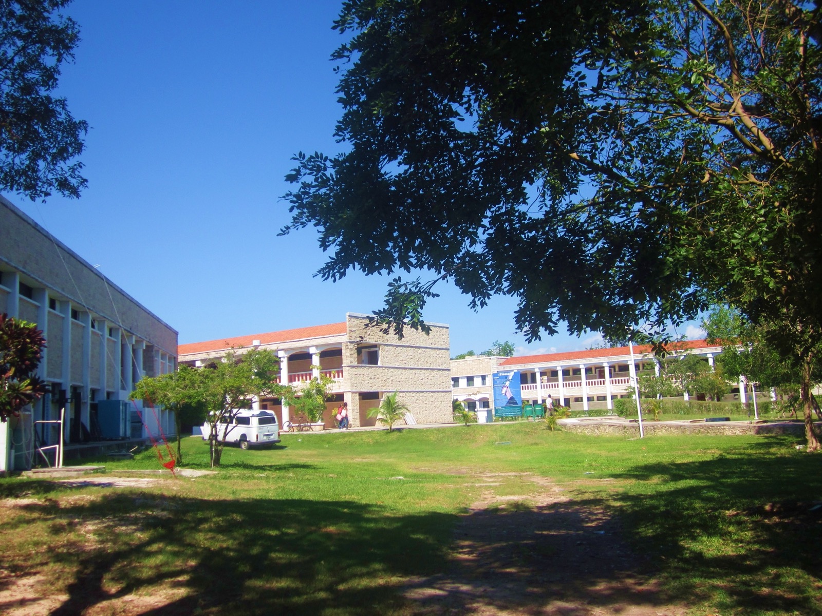Two story university buildings with red tiled roofs across a sunlit lawn and shade trees on the Universidad de Quintana Roo campus in Chetumal