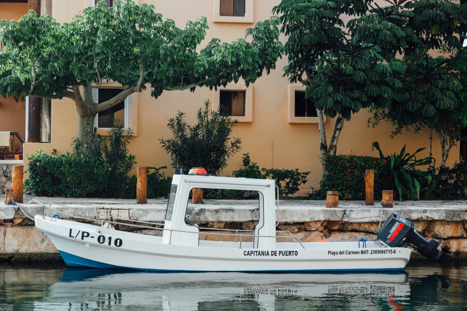 The white Capitanía de Puerto launch L/P 010 moored against the stone quay of the Puerto Aventuras marina canal, with trees and a peach apartment building behind
