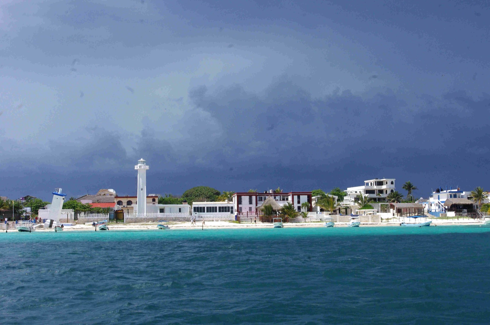 The Puerto Morelos waterfront seen from the sea under a dark storm sky, with the leaning old lighthouse, the white working lighthouse, low beachfront buildings and small boats moored off a strip of white sand