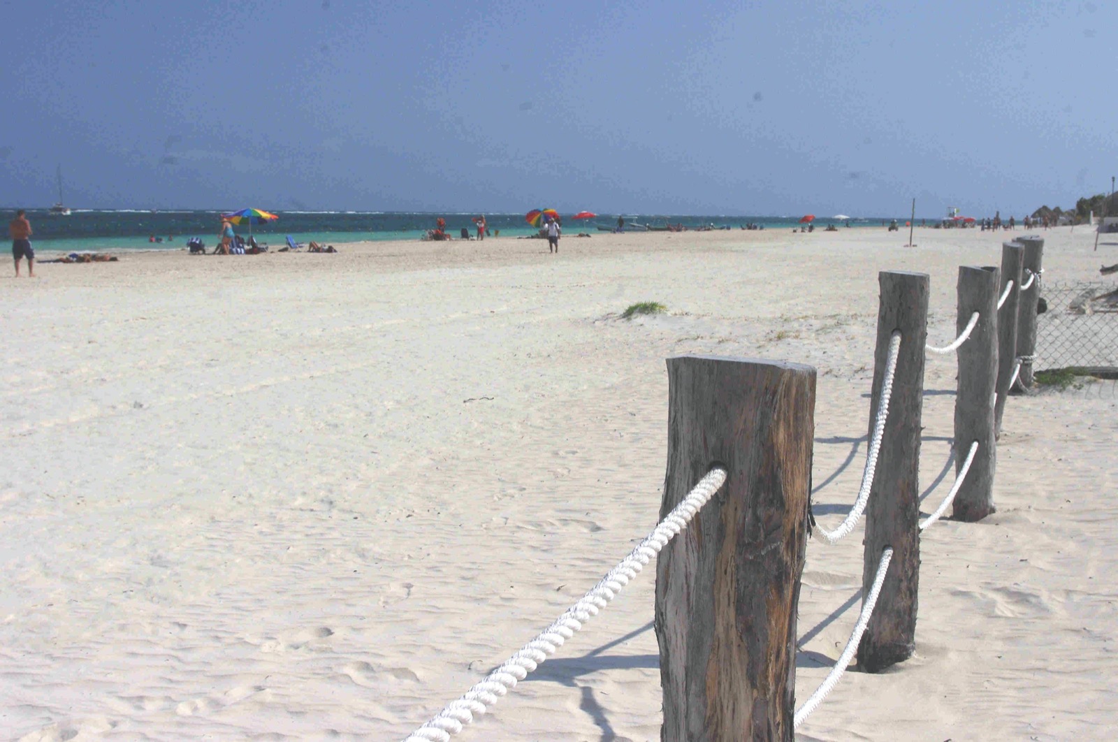 A wide white sand beach in Puerto Morelos with a line of timber posts and rope in the foreground, sunshades along the water and the reef breaking on the horizon