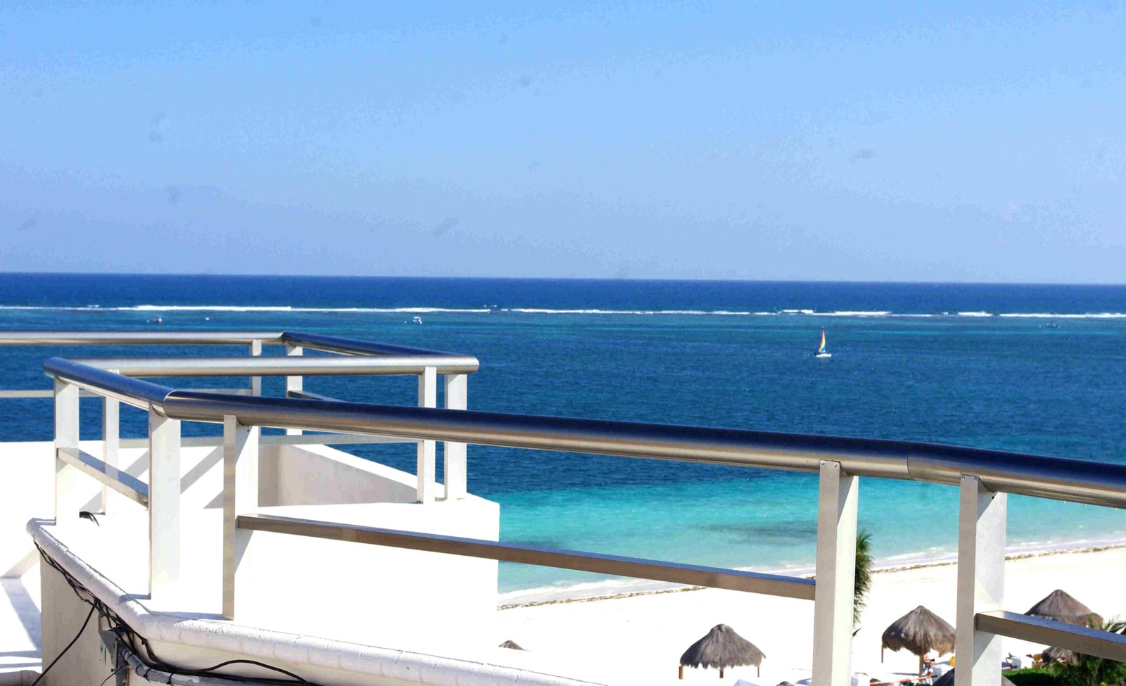 View from a white rooftop terrace in Puerto Morelos over a turquoise lagoon to the line of surf breaking on the offshore reef, with a small sailboat and palapa sunshades on the beach below