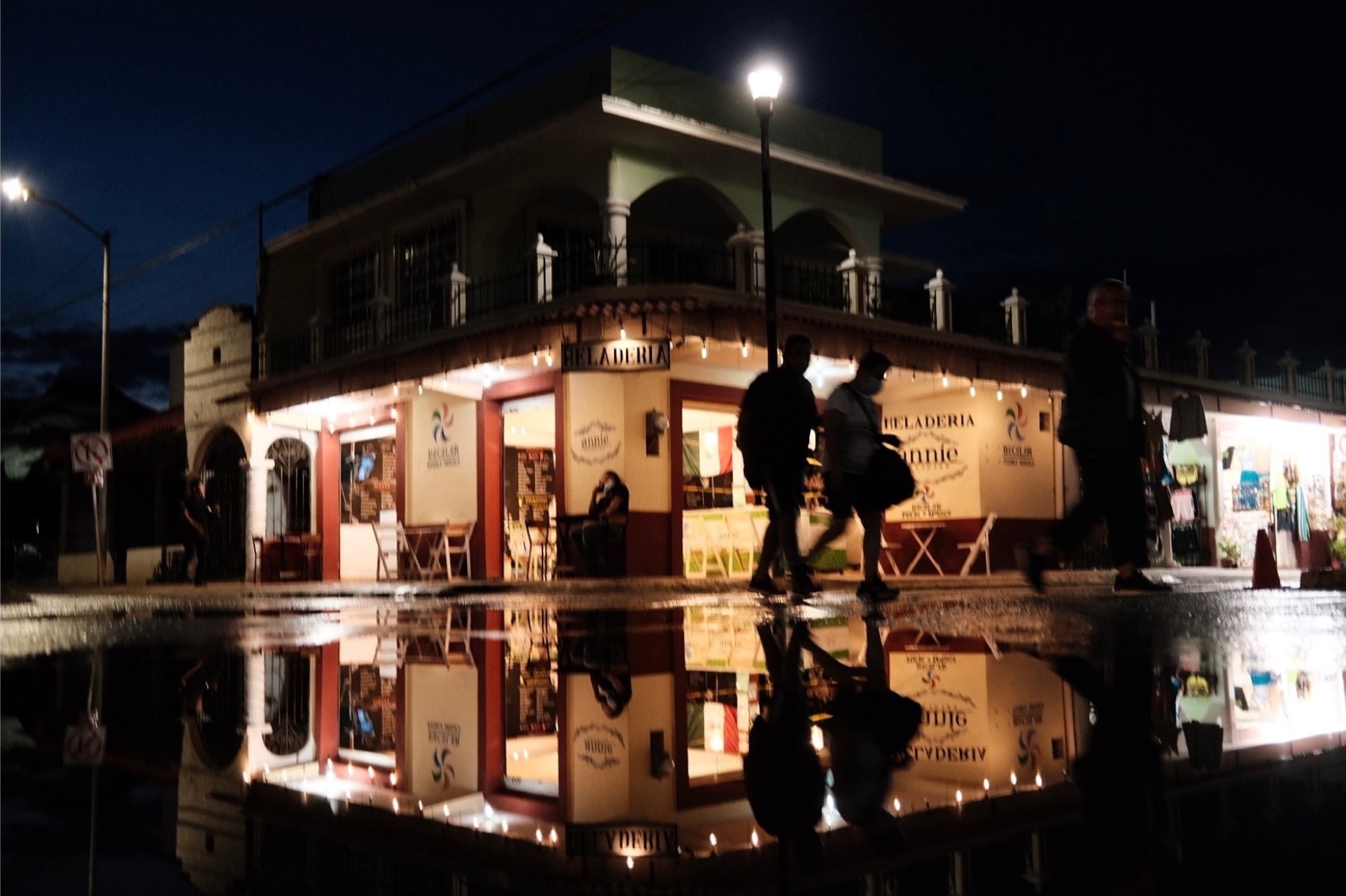 Rain water pooled on a Bacalar street at night, reflecting a lit corner ice cream shop as people walk past