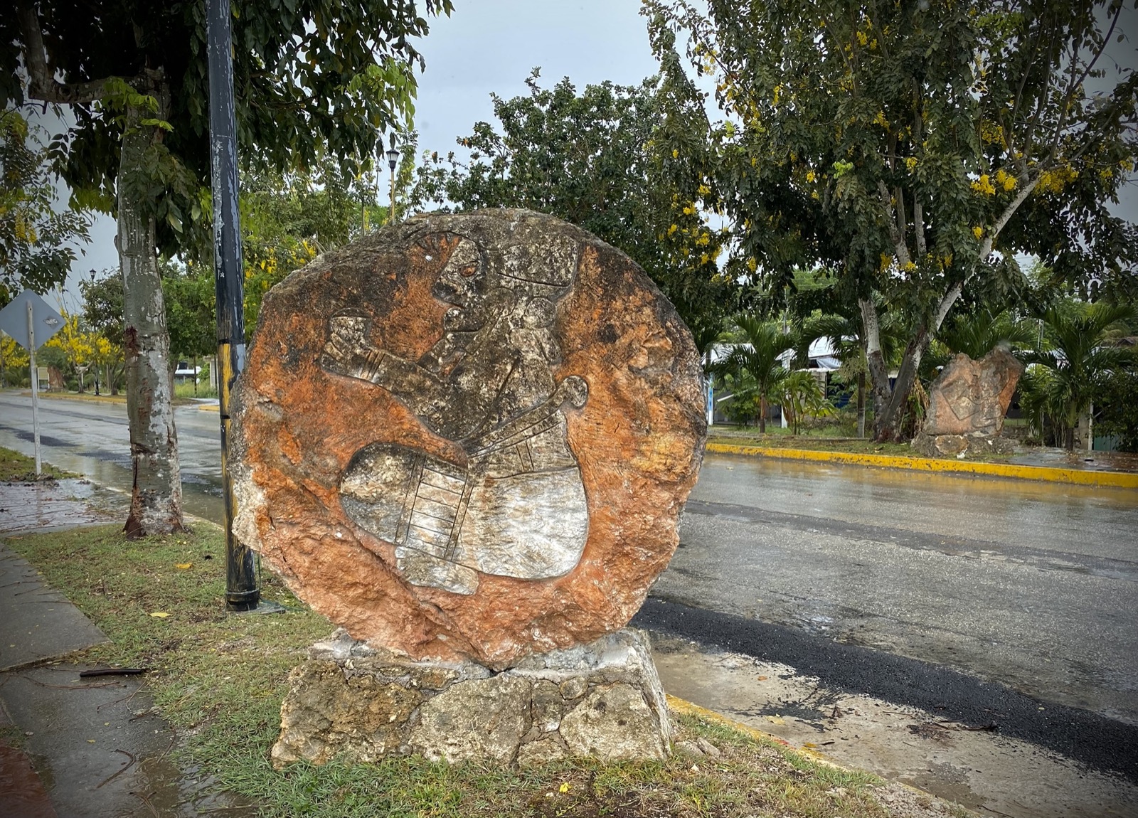 A carved stone disc beside a rain soaked avenue in Bacalar, with yellow painted curbs and flowering trees beyond