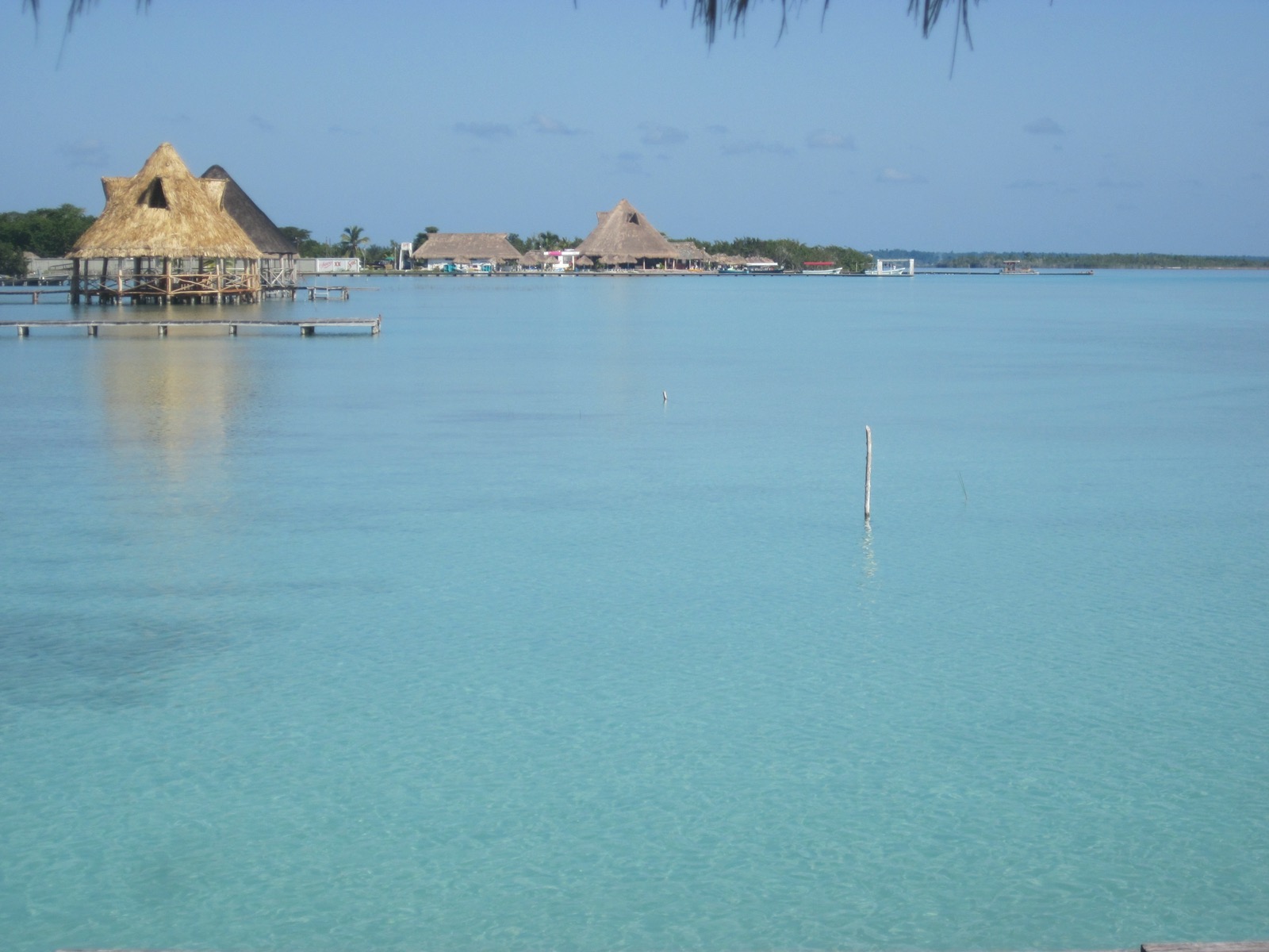 Thatched palapas and wooden piers along the shore of the Bacalar lagoon, with clear turquoise water in the foreground