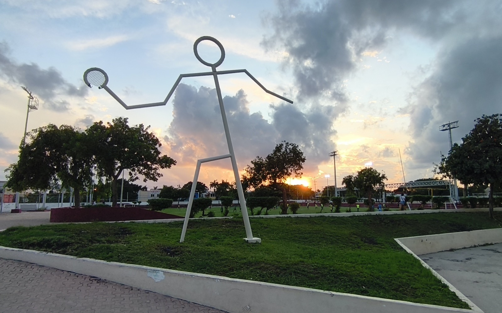 A tall white steel sculpture of a stick figure swinging a racket stands on a lawn at the Poliforum sports complex in Playa del Carmen, with trees, floodlights and a sunset sky behind