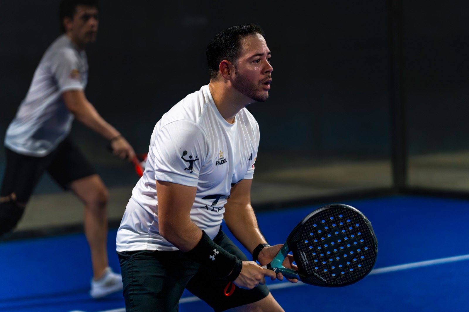 A padel player in a white shirt waits at the ready with a perforated black racket on a bright blue indoor court, his partner behind him