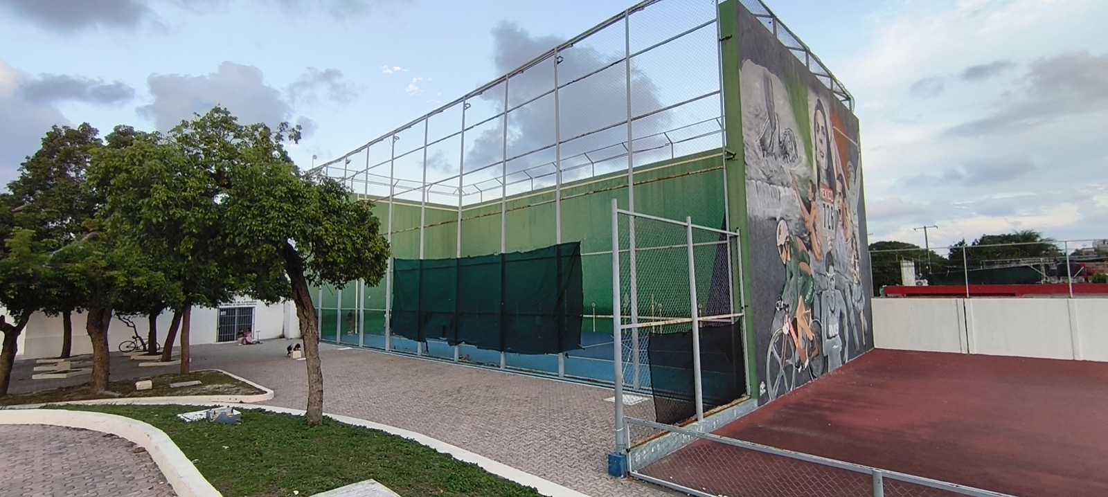 The high green walls and fencing of the frontón courts at the Poliforum sports complex in Playa del Carmen, beside a sports mural, trees and a paved walkway