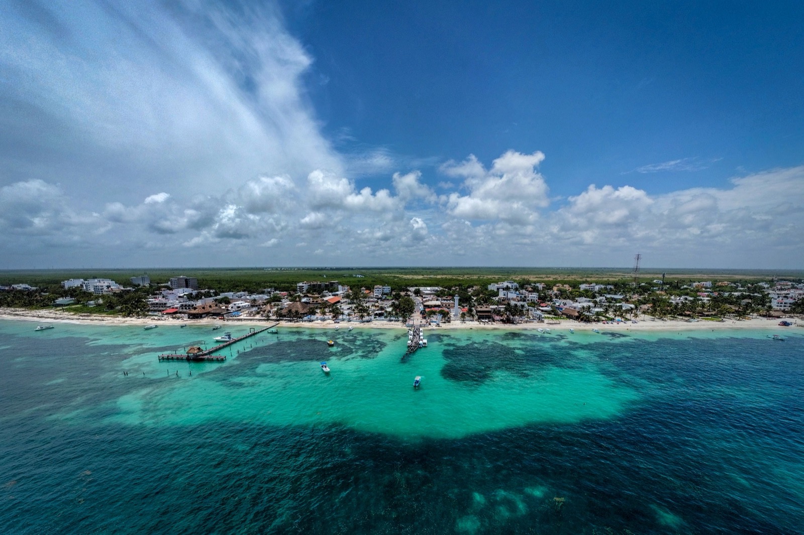 Aerial view of Puerto Morelos from the sea: turquoise reef water, two piers with small boats, a white beach and the low rooftops of the old town under a big sky