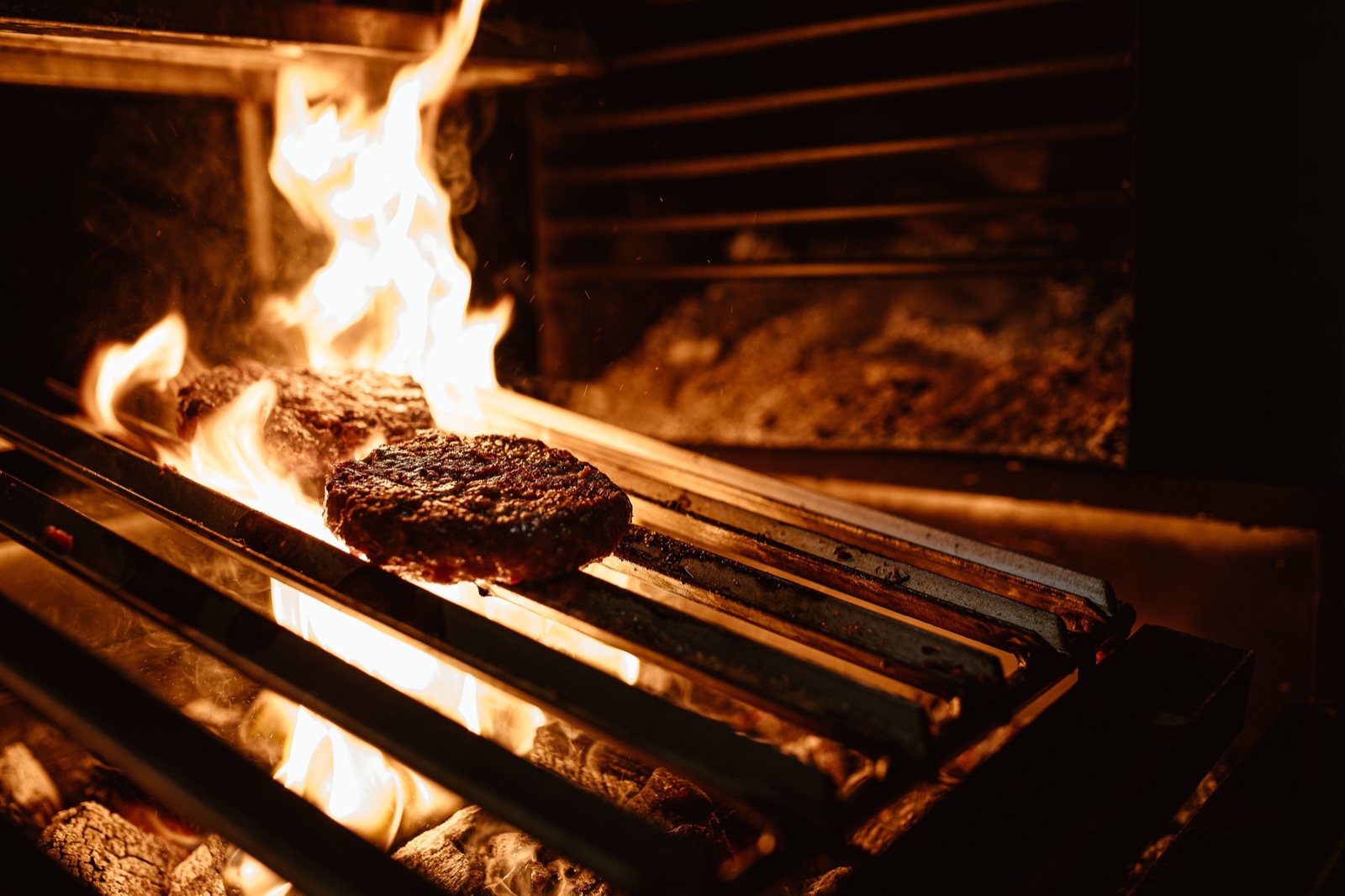 Wood and charcoal burning under a steel grill, with flames rising around two patties of meat searing on the bars