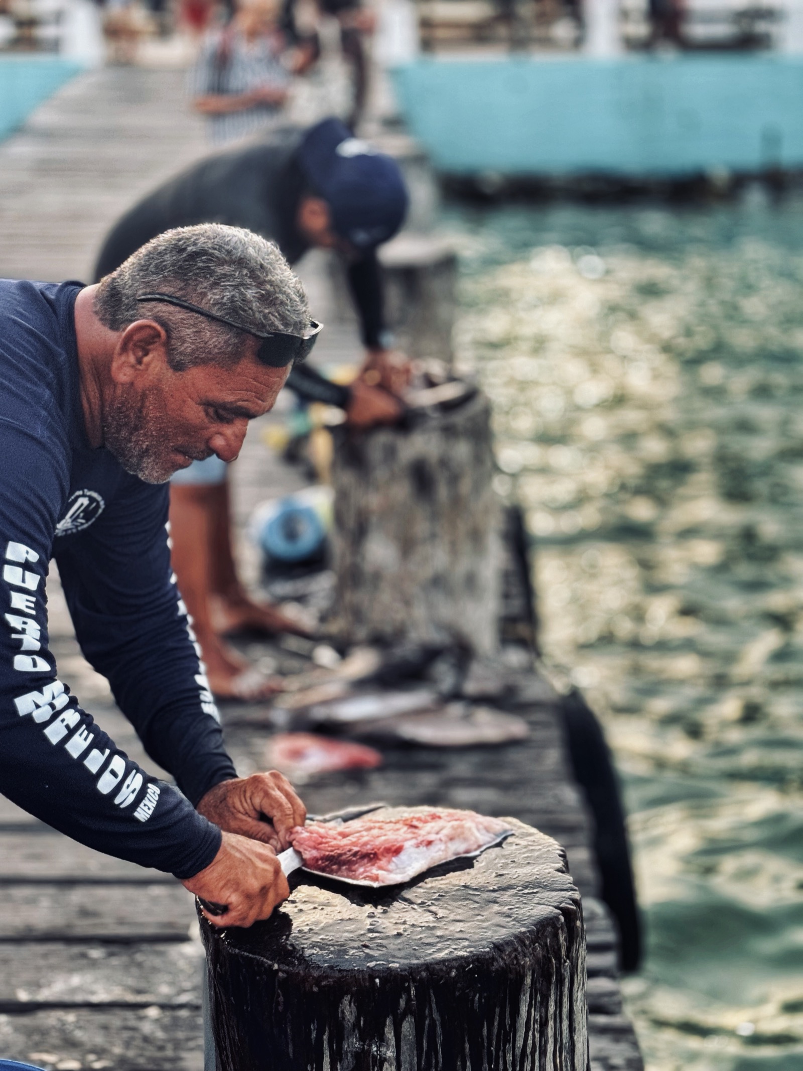 A fisherman in a Puerto Morelos shirt filleting a fresh fish on a wooden post of the town pier, with another man working further along and turquoise water beside them