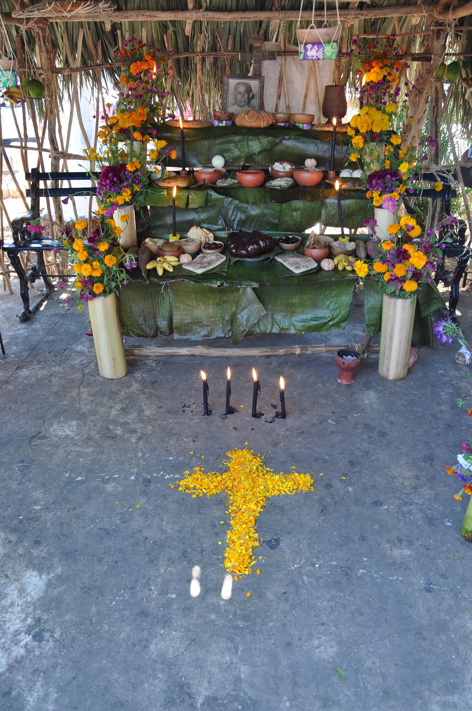 A Janal Pixán altar under a palm thatch roof, laid with bread, bowls of food, fruit and candles and framed with marigolds, with a cross of marigold petals on the ground in front