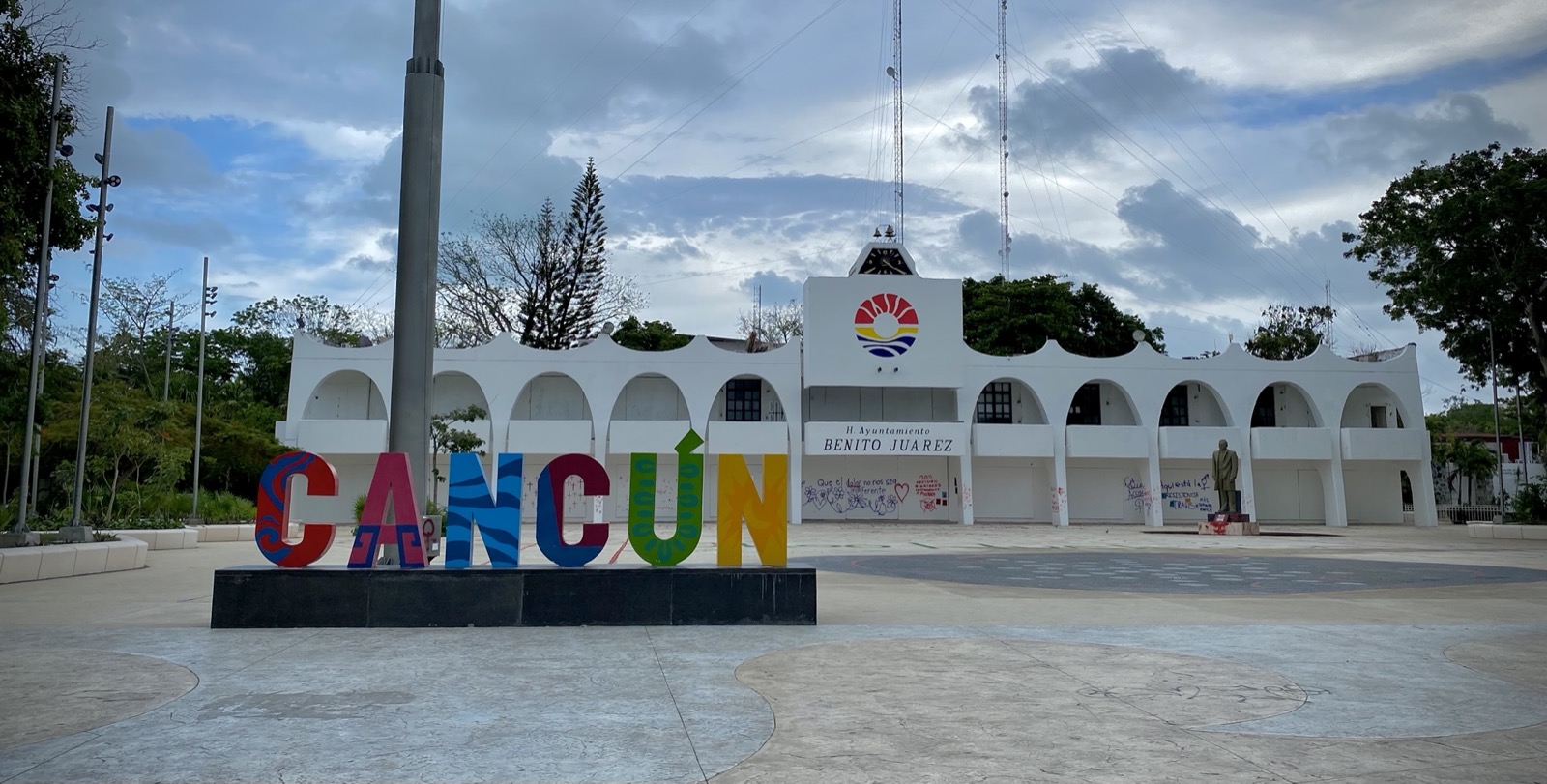 Cancún's municipal palace, the white arcaded H. Ayuntamiento Benito Juárez building, behind the colorful CANCÚN letters on its plaza under a cloudy sky