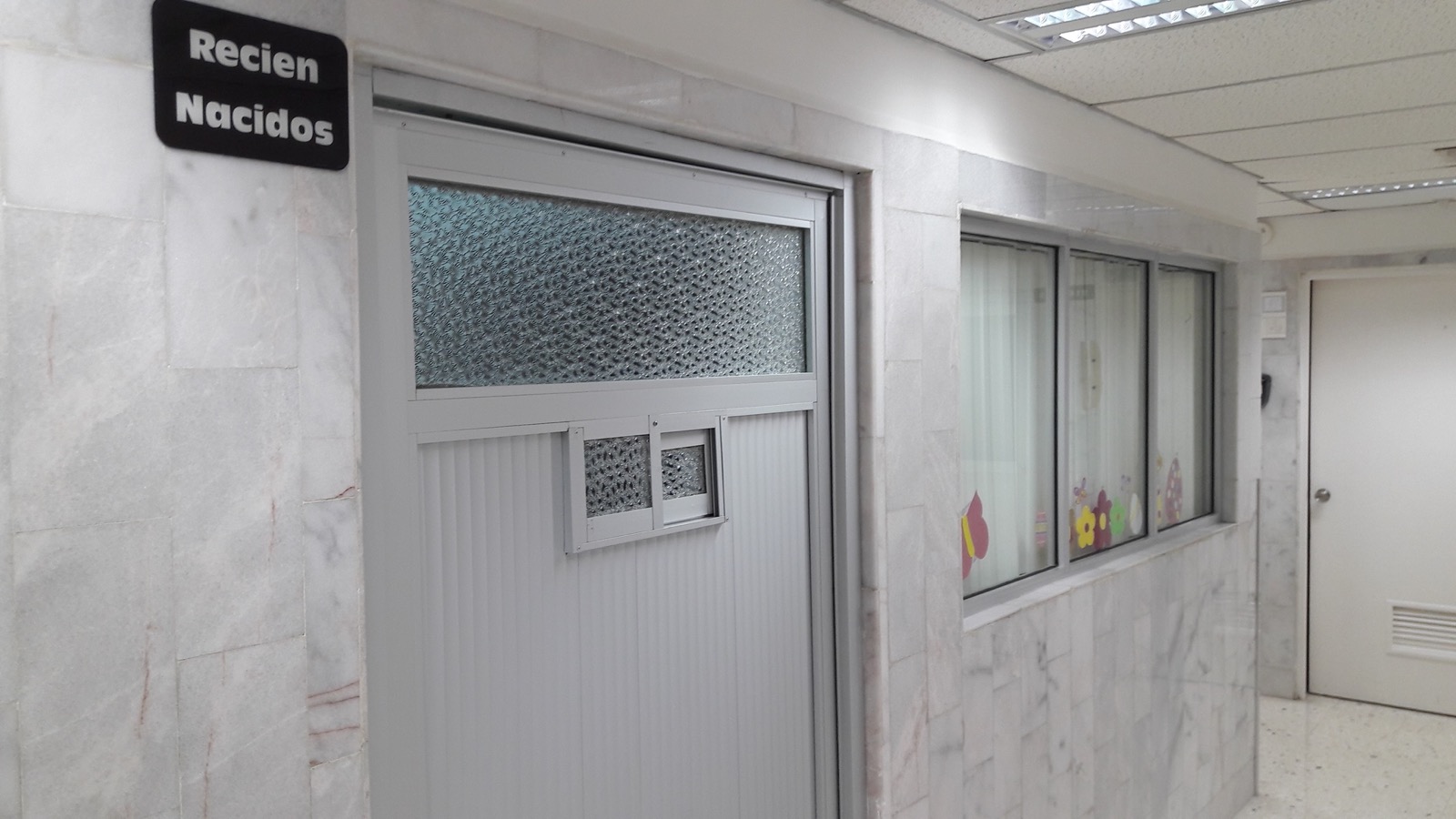A hospital corridor in Mexico with a door marked Recién Nacidos and a nursery window decorated with paper flowers
