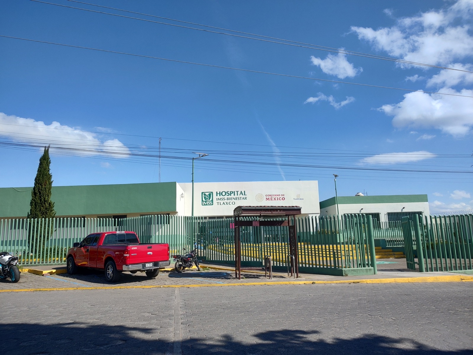 The single story green and white IMSS-Bienestar hospital in Tlaxco, Tlaxcala, behind a green railing fence under a blue sky