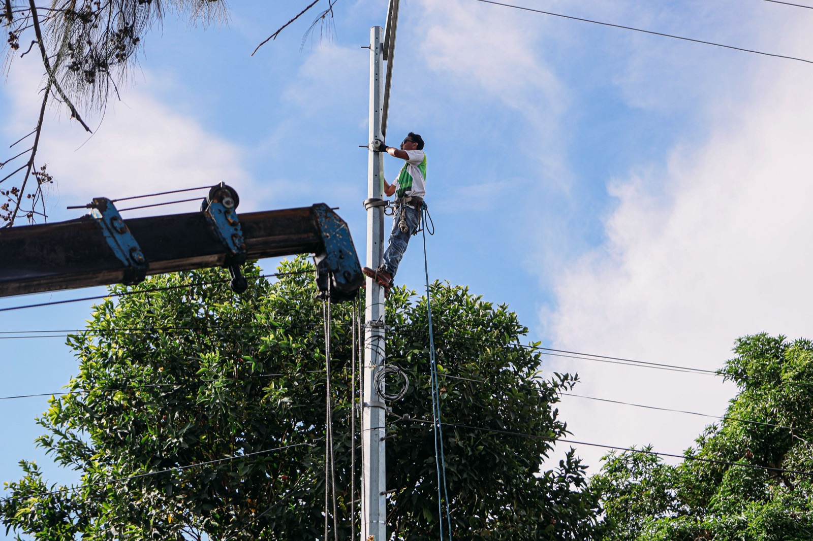 A utility worker in a safety harness climbing a tall metal pole, with a crane arm beside him and leafy trees and blue sky behind